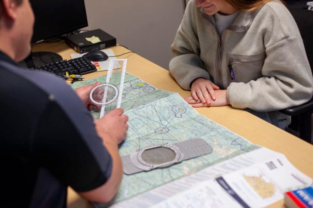 An instructor and a student at the briefing table, working across a sectional chart with a plotter between them.